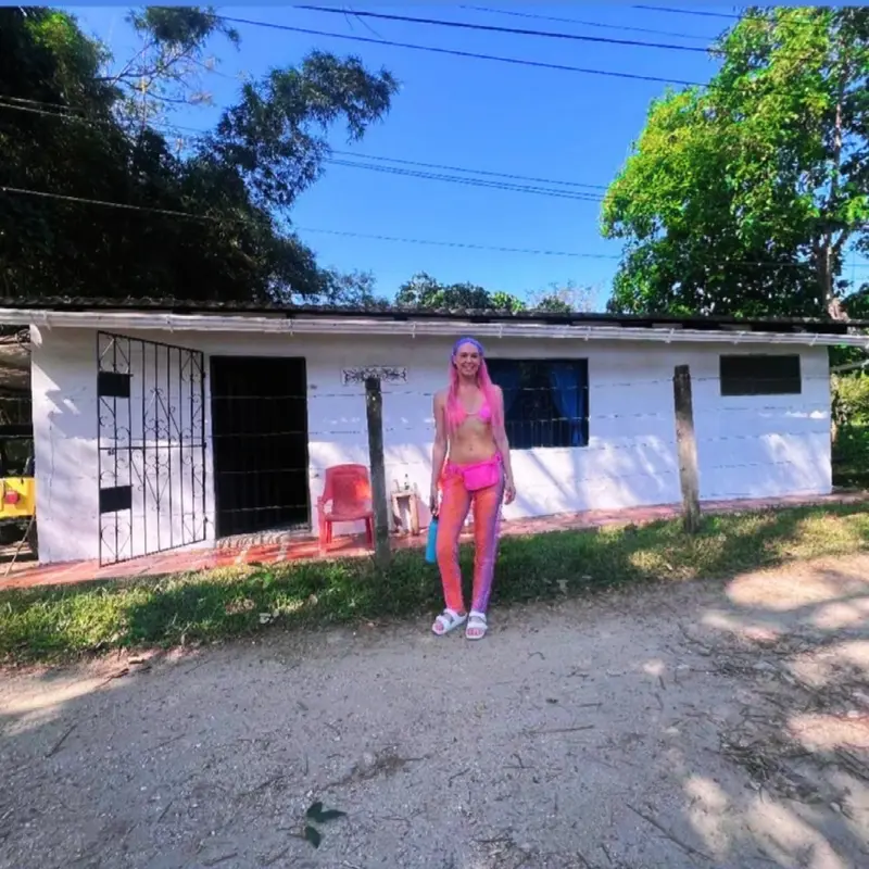 A woman in pink posing on the dirt lane in front of a white single-storey house with barred windows