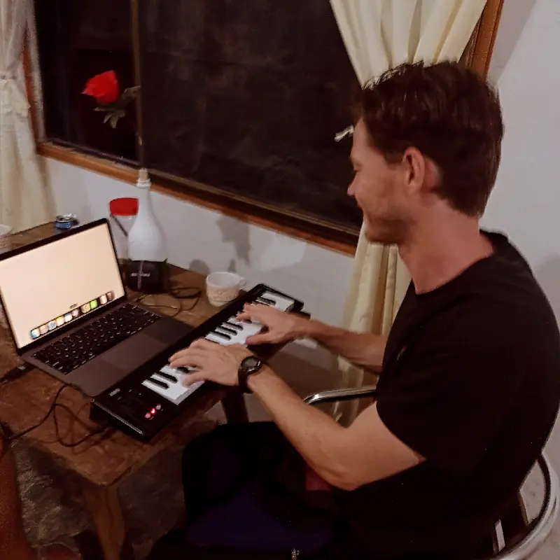 A man playing a small keyboard connected to a laptop at a wooden desk, a red rose in a bottle beside him