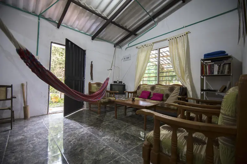 The living area with wooden armchairs, cushions, a bookshelf and a hammock, light coming through curtained windows