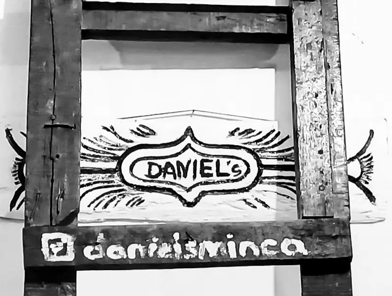 Hand-painted black-and-white sign with the word DANIEL’S inside a winged emblem and the handle @danielsminca on a wooden frame