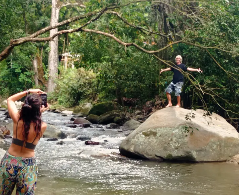 A white-haired man standing arms wide on a huge boulder in a rushing river while a woman photographs him