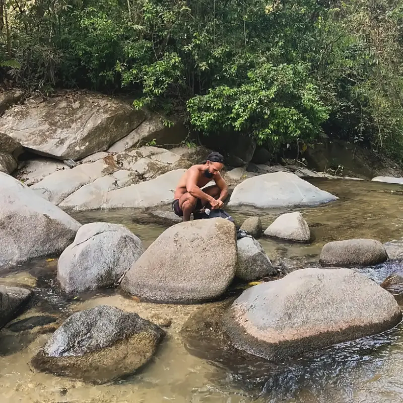 A man crouching on a boulder in a clear, shallow mountain river surrounded by big granite stones