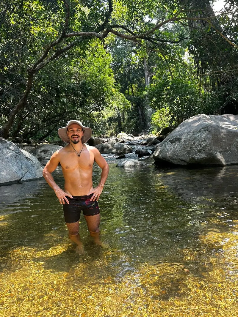 A man in a hat standing knee-deep in a clear, sunlit jungle river